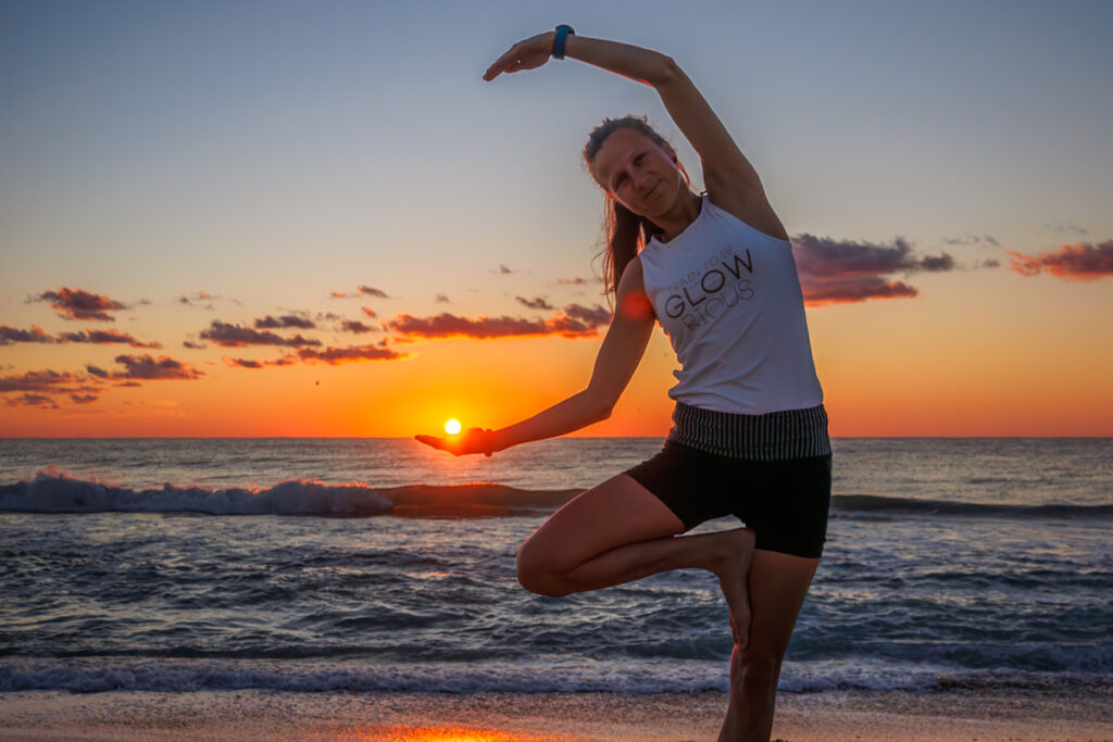 Yogalehrerin vor dem Meer zum Sonnenuntergang hält optisch den Sonnenball in ihrer Hand